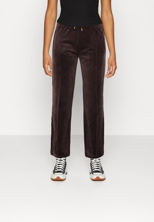 Person, der bærer mørkebrune fløjlsbukser med lodrette sømme, sort tanktop og hvide-sorte sneakers, står mod en ensfarvet baggrund.