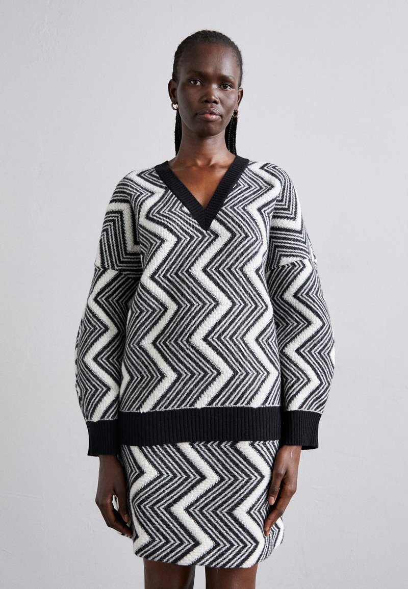 Knitted sweater dress in black and white with zigzag pattern, featuring a V-neck, long sleeves, and ribbed cuffs and bottom hem.