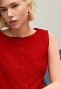 Haut sans manches texturé rouge avec un col rond et cinq boutons assortis sur le devant. Tissu lisse, coupe décontractée.