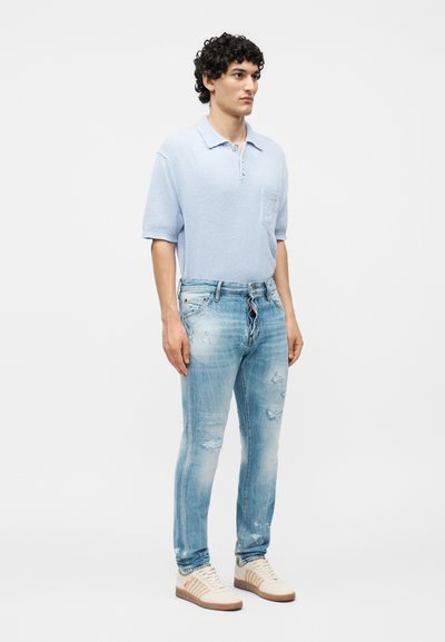 Νέος άνδρας με σγουρά μαλλιά φορά ένα ανοιχτό μπλε πόλο με κοντά μανίκια, ξεβαμμένα ανοιχτό μπλε τζιν και λευκά αθλητικά παπούτσια, στέκεται σε μονόχρωμο φόντο.
