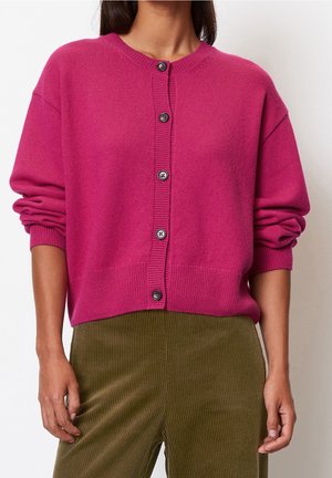 Kobieta ubrana w jasnoróżowy sweter na guziki z prążkowanymi mankietami oraz oliwkowozielone prążkowane spodnie, stojąca na tle białego tła.