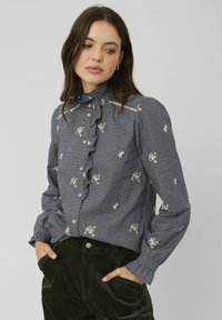 Blouse à carreaux noir et blanc avec un devant froncé, broderies florales et col montant. Manches longues avec poignets à boutons. Associée à un pantalon vert foncé.