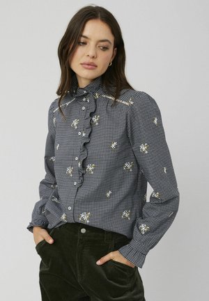 Blouse à carreaux noir et blanc avec un devant froncé, broderies florales et col montant. Manches longues avec poignets à boutons. Associée à un pantalon vert foncé.
