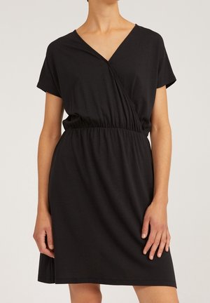 Mujer con un vestido negro cruzado de manga corta con cintura elástica, de pie con una mano relajada y la otra sujetando el dobladillo del vestido.