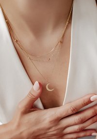 Collana con strati d'oro che presenta un pendente a forma di luna crescente, catene delicate e accenti sferici, esposta su una pelle liscia.