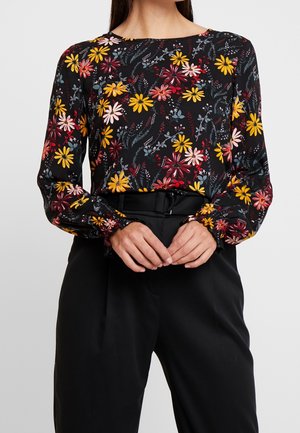 Schwarzes Langarmshirt mit gelbem, rotem und pinkfarbenem Blumenmuster, kombiniert mit hoch taillierten schwarzen Hosen und einem schwarzen Gürtel.