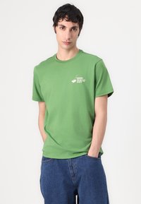 T-shirt de algodão verde com gola redonda, mangas curtas e uma impressão gráfica branca no lado esquerdo do peito. Acompanhado de jeans de ganga azuis.