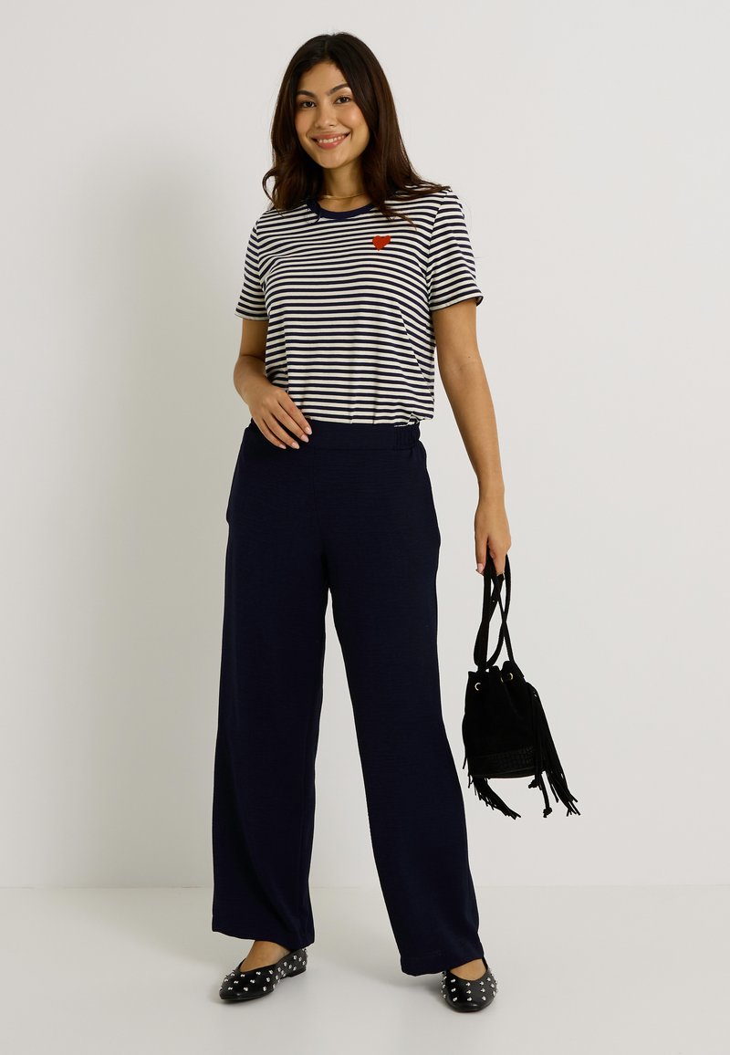 T-shirt rayé bleu marine et blanc avec détail de cœur rouge, associé à un pantalon large bleu marine et un sac noir avec des accents de franges. Chaussures plates noires avec clous.