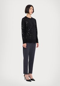 Schwarzer Pullover mit Pailletten-Punkte-Muster, kombiniert mit dunkelgrauen, maßgeschneiderten Hosen und schwarzen spitzen High Heels. Schlichte, strukturierte Gestaltung.