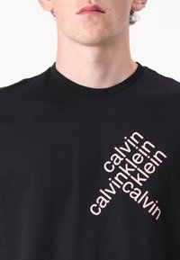 Zwarte katoenen t-shirt met ronde hals, met herhaaldelijk roze tekst die "calvin klein" in een hoek leest. Glad textuur, relaxed fit.