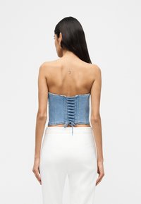 Γυναίκα με μακριά σκούρα μαλλιά που φοράει κορσέ από τζιν σε ανοιχτό μπλε χρώμα με κορδόνια και λευκό παντελόνι, εμφανίζοντας ένα μικρό τατουάζ σε σχήμα ημισελήνου στον πάνω μέρος της πλάτης της.