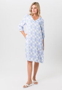 Hellblau gemustertes Kleid mit V-Ausschnitt, drei Viertelärmeln und zwei seitlichen Taschen. Der Stoff hat eine glatte Textur und ein geometrisches Design.