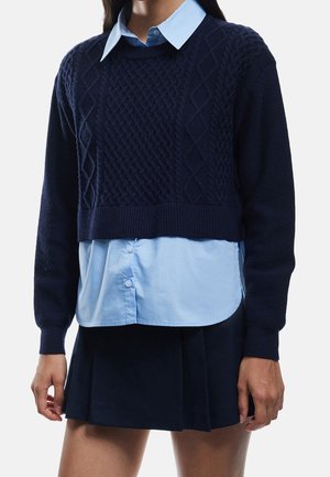 Mujer que lleva un jersey corto azul marino de punto con trenzas sobre una camisa azul clara con botones y una falda de tablas azul marino.