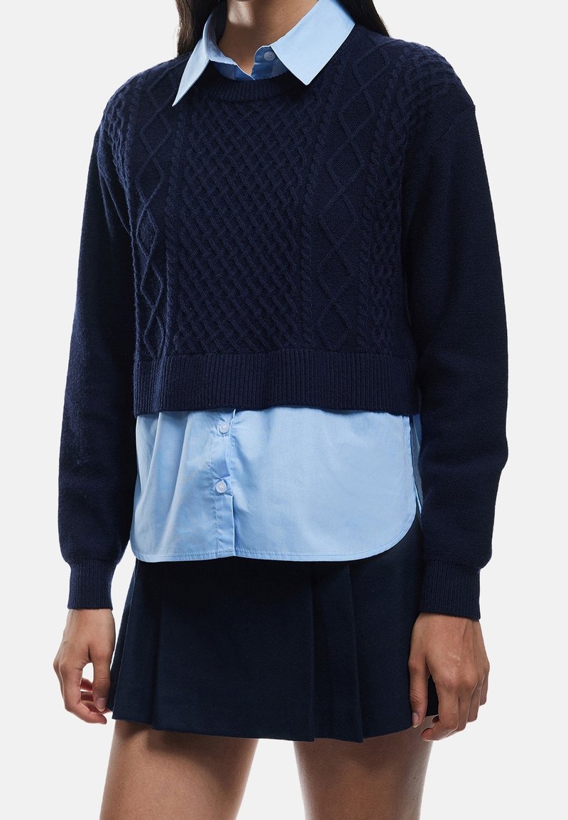 Femme portant un pull court bleu marine en maille torsadée par-dessus une chemise bleu clair boutonnée et une jupe plissée bleu marine.