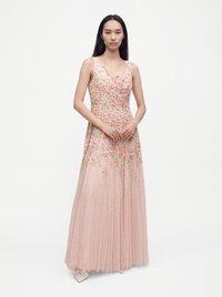 Femme portant une robe rose sans manches avec broderies florales et une jupe fluide, debout devant un fond blanc.