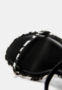 Sandale noire avec une semelle texturée, dotée d'une sangle à motif noir et blanc. Le matériau a une finition lisse avec des accents duveteux remarquables.
