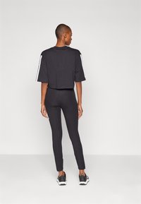 T-shirt preto cropped com riscas brancas nas mangas, combinado com leggings pretas. Modelo usa sapatilhas de desporto pretas. Tecido suave, design ajustado.