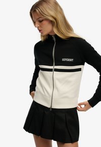 Czarna i biała bluza na zamek z fakturowanej tkaniny, z srebrnym zamkiem i logo „SUPERDRY” na piersi. Poniżej plisowana czarna spódnica.