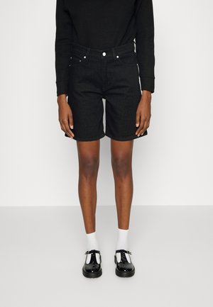 Personne portant un haut noir à manches longues, un short en jean noir longueur genou, des chaussettes blanches aux chevilles et des chaussures Mary Jane en cuir verni noir.
