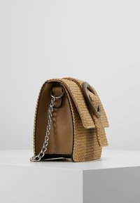 Malá hnědá tkaná kabelka s velkou kulatou sponou na klopě a krátkým stříbrným řetízkovým popruhem, umístěná na bílé ploše na pozadí v jednobarevné šedé.