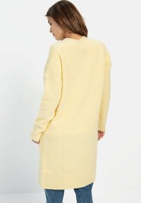 Lange, gele gebreide cardigan met een V-hals en een losse pasvorm. Heeft lange mouwen en een licht gestructureerd oppervlak. Gecombineerd met jeans.