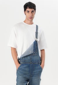 Denim tuinbroek met een bies aan de voorkant heeft een lichte wasaccent, verstelbare schouderbanden en een zak. Gecombineerd met een basic wit T-shirt.