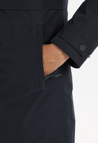 Dettaglio di giacca impermeabile blu navy con tasca laterale, polsino elastico e accento di bottone visibile. Tessuto testurizzato con un design elegante.