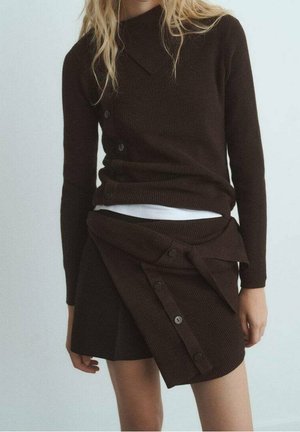 Femme portant un haut en maille marron foncé à manches longues et une jupe asymétrique boutonnée assortie par-dessus un sous-pull blanc, debout devant un fond uni.