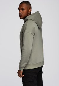 Nuori mies, jolla on päällä vaaleanharmaa huppari ja mustat cargo-housut, seisoo sivuttain yksinkertaista harmaata taustaa vasten.