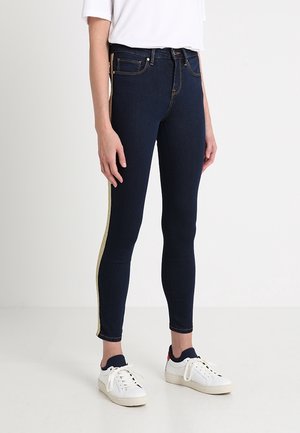 Femme portant un jean skinny foncé avec une bande latérale dorée, des baskets blanches et un haut blanc à manches courtes, debout sur un sol blanc.