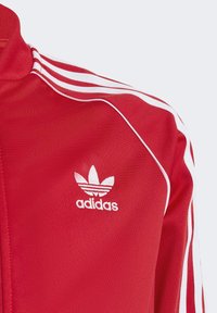 Czerwona kurtka dresowa Adidas z białymi paskami i logo. Posiada wysoki kołnierz oraz teksturowaną tkaninę, podkreślającą sportowe elementy designu.
