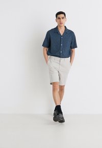 Camisa de manga corta azul marino con un patrón sutil, combinada con pantalones cortos color beige claro. El modelo lleva calcetines negros y zapatos oscuros.