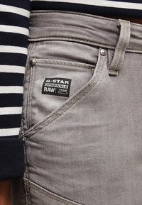 Jeans de mezclilla G-Star Raw en gris que presentan una etiqueta distintiva, un bolsillo delantero y un diseño de costura plana y reforzada. Camiseta de rayas en blanco y negro a la vista.