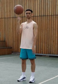 Sportlicher Mann in einem hellrosa ärmellosen Oberteil und türkisfarbenen Shorts, der einen Basketball auf einem Finger dreht. Der Hintergrund besteht aus Holzpaneelen.