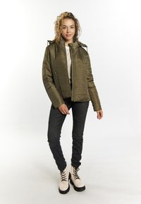 Giacca isolata verde oliva con zip frontale e cappuccio, abbinata a jeans neri slim e stivaletti crema con suole e lacci neri.