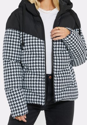 Femme portant une doudoune à motif pied-de-poule noir et blanc avec une capuche noire et une fermeture éclair, par-dessus une chemise blanche et un pantalon noir.