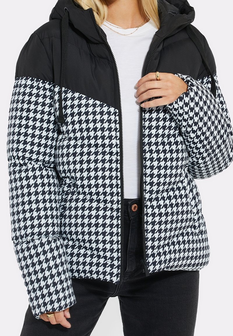 Femme portant une doudoune à motif pied-de-poule noir et blanc avec une capuche noire et une fermeture éclair, par-dessus une chemise blanche et un pantalon noir.