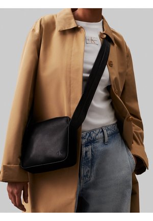 Černá krosbody taška s hladkou texturou, s jemným logem. Nosená s béžovým kabátem a světlými džíny, což ukazuje na ležérní styl.