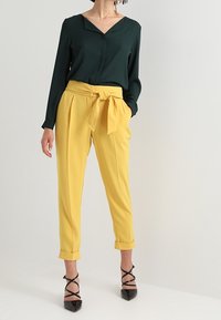 Femme portant un chemisier à manches longues vert foncé, un pantalon taille haute jaune style paperbag avec ceinture nouée, et des talons noirs à lanières avec bout pointu.