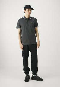 Polo grigio con dettagli bianchi al colletto e piccolo logo giallo, abbinato a pantaloni felpati neri e sneakers nere, in piedi davanti a uno sfondo chiaro.