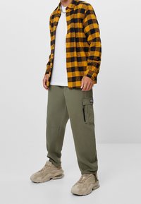 Geel en zwart geruit flanellen shirt over een witte T-shirt; olijfgroene cargo broek met een ritssluiting; tan sneakers met een gestructureerde bovenkant.