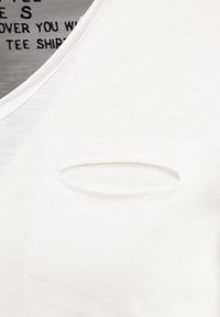 Camiseta blanca de algodón con escote redondeado, que presenta un exclusivo bolsillo ovalado recortado y una textura suave, exhibiendo detalles de costura sutiles.