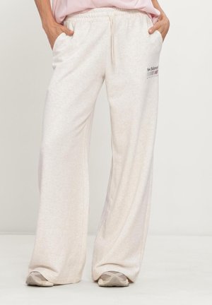 Persona con pantalones deportivos sueltos beige de New Balance con cintura elástica, manos en los bolsillos, combinados con zapatillas beige y blancas.