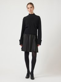 Kvinna klädd i svart tröja med långa ärmar och vida manschetter, svart skinnkjol, svarta strumpbyxor och svarta klackar, stående mot en ljus enfärgad bakgrund.