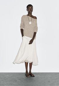 Beige gebreide trui met off-shoulder ontwerp, gecombineerd met een vloeiende witte enkellange rok. Model draagt slippers en een gestructureerde ketting.