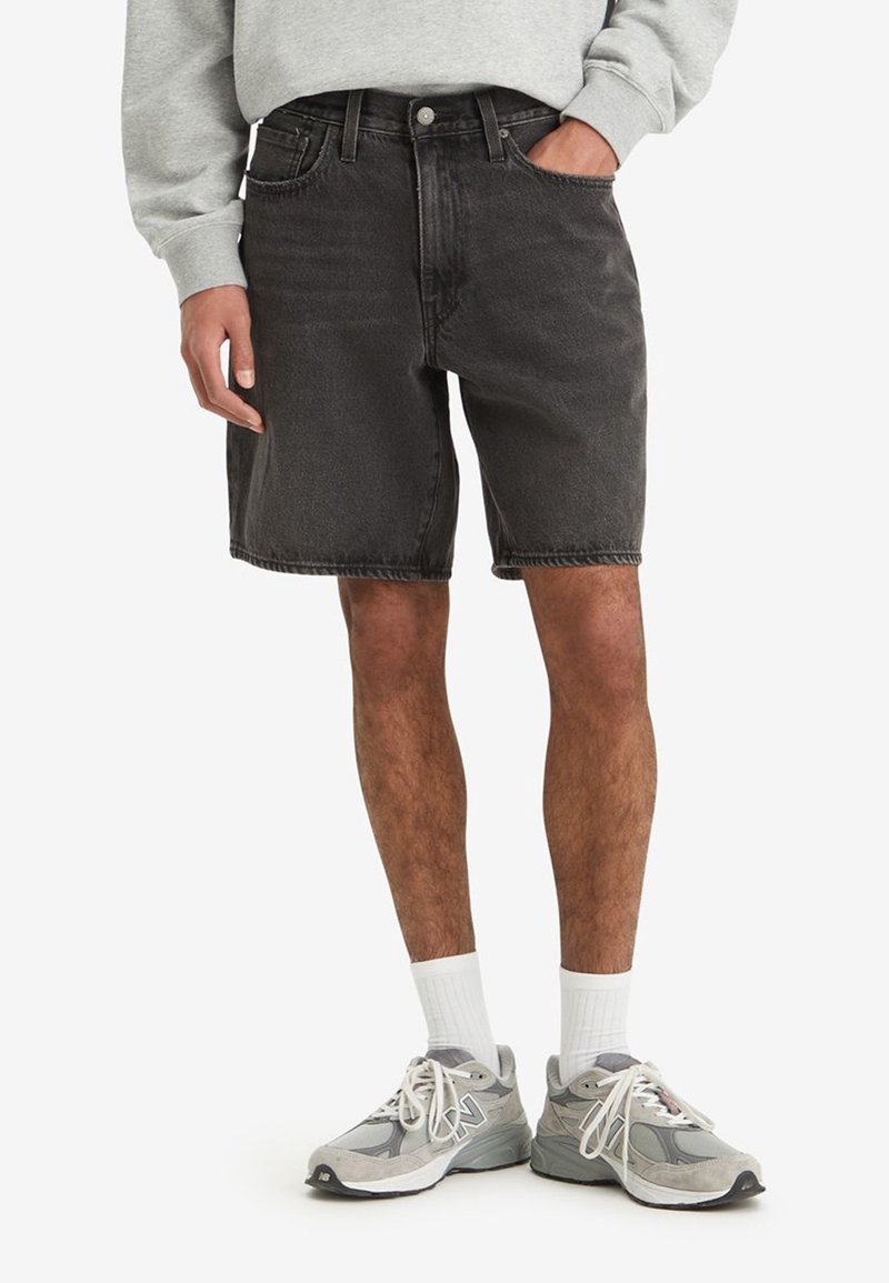 Homme portant un short en jean noir, un sweat-shirt gris clair, des chaussettes blanches montantes et des baskets de sport grises avec des lacets.