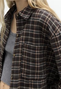 Camisa de franela de cuadros negros y marrones con cuello extendido, cierre de botones y un bolsillo en el pecho. La tela tiene una textura suave.