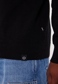 Black knitted pullover with ribbed cuffs and hem, featuring a small black label with a white diamond logo. Paired with light blue denim.