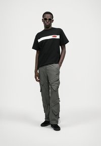 Camiseta preta com faixa branca e logotipo no peito, combinada com calças cargo largas em tom de oliva, com vários bolsos. Sapatos pretos.