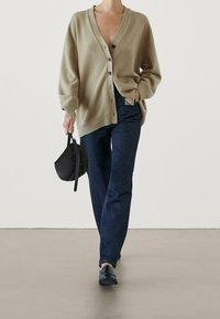 Beige wool cardigan with a deep V-neck and black buttons, paired with dark blue jeans and a black round handbag, worn with black shoes.